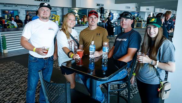 A group of five NASCAR fans stands around a high-top table inside the Newton Club at Iowa Speedway, enjoying drinks in the climate-controlled hospitality lounge before race activities. A concession area and other guests are visible in the background.